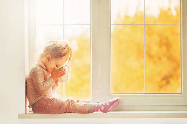 Menina bonito criança no pijama sentado à janela grande jogando sorrindo desfrutar de casa. Copo de cacau em pé na janela. Outono Temporada Interior Família Indústria têxtil Férias Pessoas conceito
 - Foto, Imagem