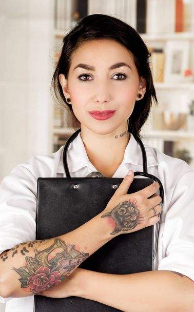 Beautiful tattooed young doctor hugging a black binder in office background - 写真・画像