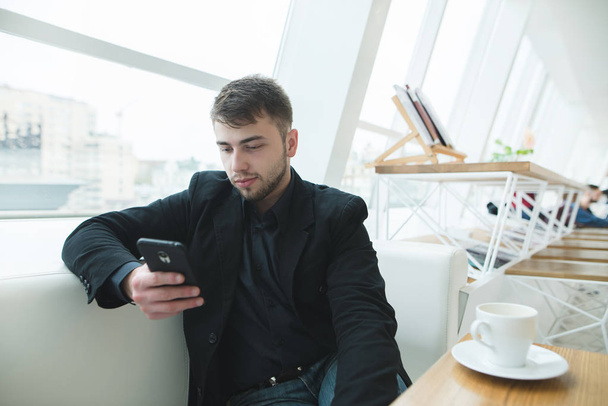 Un giovane uomo d'affari scrive un messaggio sul suo smartphone mentre è seduto in un elegante ristorante vicino alla finestra. Pausa caffè nel caffè leggero
. - Foto, immagini