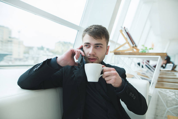 The man is sitting by the window and talking on the phone with a cup of hot coffee in his hands. Lunch in a stylish cafe with a modern design. Coffee break. - Photo, Image