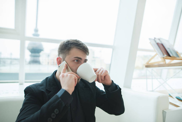 Habla por teléfono en un café para tomar una taza de café. Un hombre de traje se sienta en un elegante restaurante moderno y bebe café durante una conversación telefónica
. - Foto, Imagen