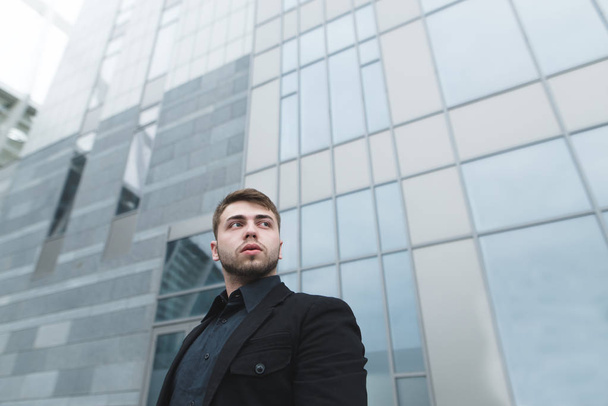 Молодой бизнесмен с бородой и в черном костюме стоит на фоне современной светлой архитектуры и смотрит вверх и вниз. Бизнес-портрет
. - Фото, изображение