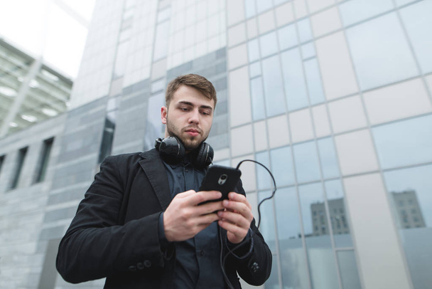 Straßenporträt eines ernsten Mannes mit Kopfhörern auf dem Hals, der vor dem Hintergrund der urbanen Landschaft Musik auf seinem Handy auswählt. - Foto, Bild