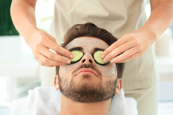 Beautician putting fresh cucumber slices on young man's face in spa salon - 写真・画像