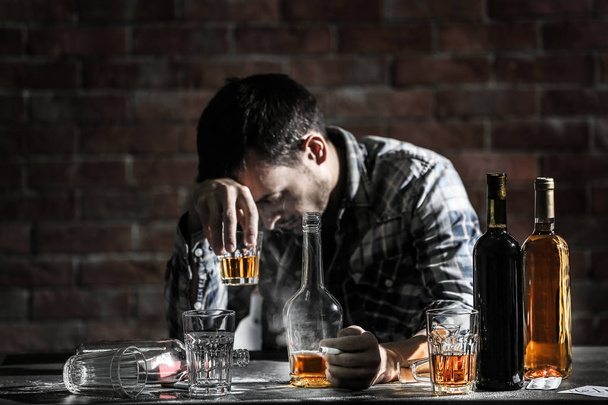 Drunk man with glass and bottles of alcohol drink sitting at table on brick wall background - Photo, Image