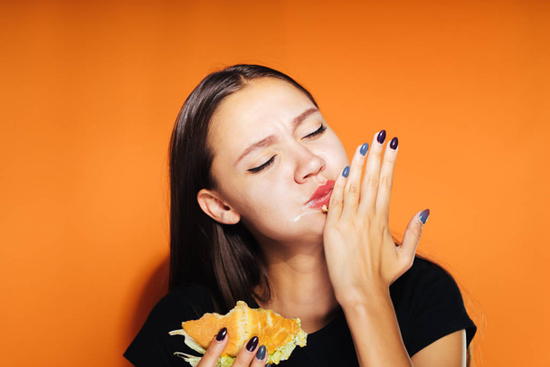 girl eagerly eats an appetizing burger and licks her hands on an orange background, isolated - Foto, imagen