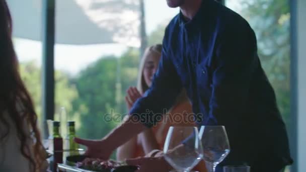 Man bringing food on table.Four caucasian friends people mediterranean italian salad,meat steak and bread lunch or dinner. Summer party at home in modern house 4k handheld video - Footage, Video