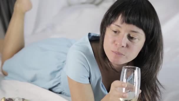 lonely young attractive woman lies on a bed in a bedroom, drinks champagne - Filmati, video