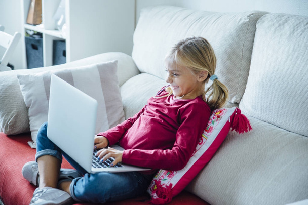 glückliches kleines Mädchen auf dem Sofa liegend mit Laptop im Wohnzimmer - Foto, Bild