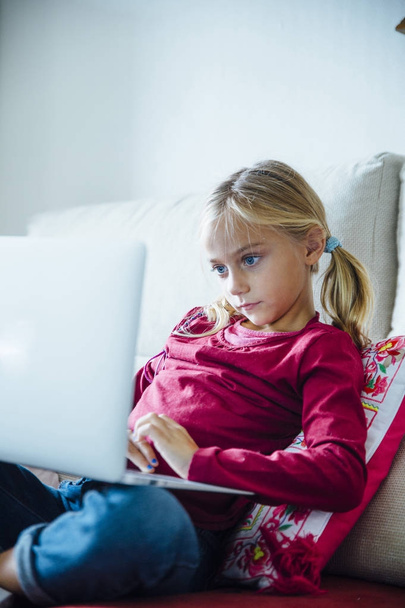 Happy  little girl lying on the sofa with laptop in the living r - Фото, изображение