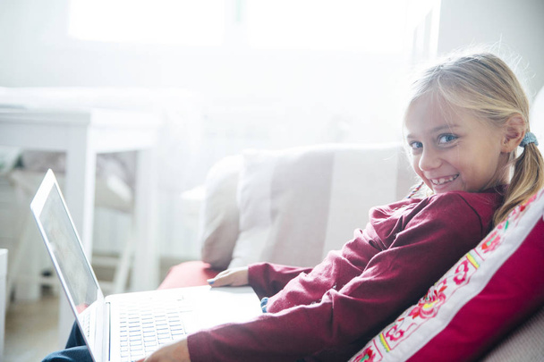 Ευτυχισμένη κοριτσάκι ξαπλωμένη στον καναπέ με το laptop στο καθιστικό με συρόμενη - Φωτογραφία, εικόνα