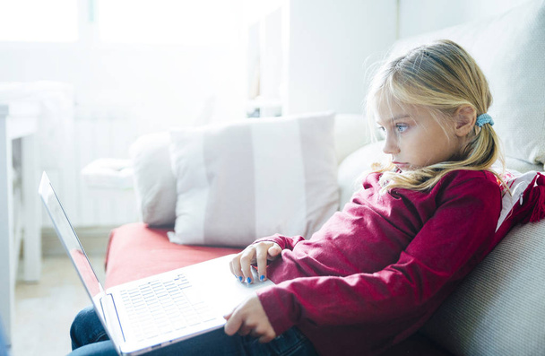 Счастливая маленькая девочка лежит на диване с ноутбуком в гостиной
 - Фото, изображение