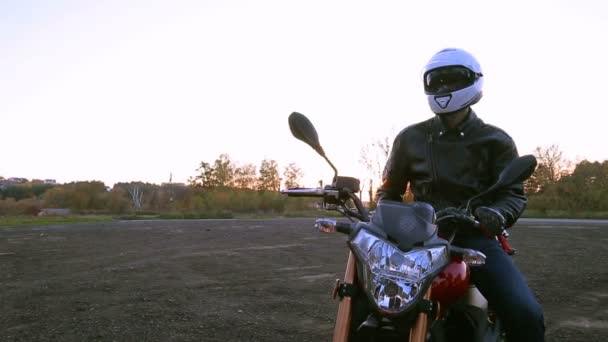 Mladý muž v černé kožené bundě a bílá helma sedí na motorce zapne světla před cestou na podzimní západ slunce - Záběry, video