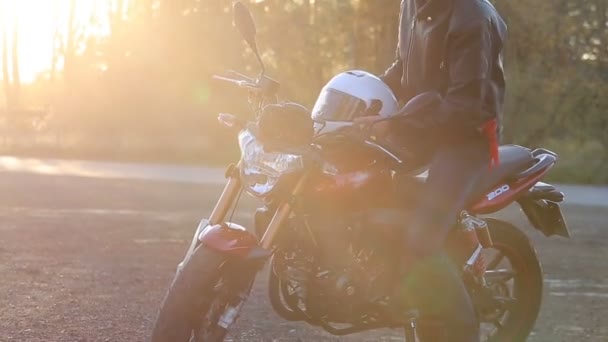 ein junger Mann in schwarzer Lederjacke mit weißem Helm steigt zu seinem Motorrad, setzt sich darauf und zeigt vor der Fahrt bei Herbstsonnenuntergang den Mittelfinger in die Kamera. - Filmmaterial, Video