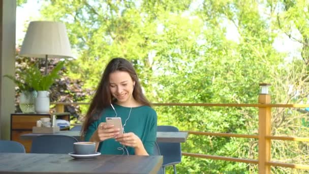 Mujer joven con teléfono inteligente mientras está sentada sola en la cafetería durante el día cálido. Atractiva hembra con linda sonrisa mensaje de escritura con teléfono móvil mientras descansa en la cafetería
 - Metraje, vídeo