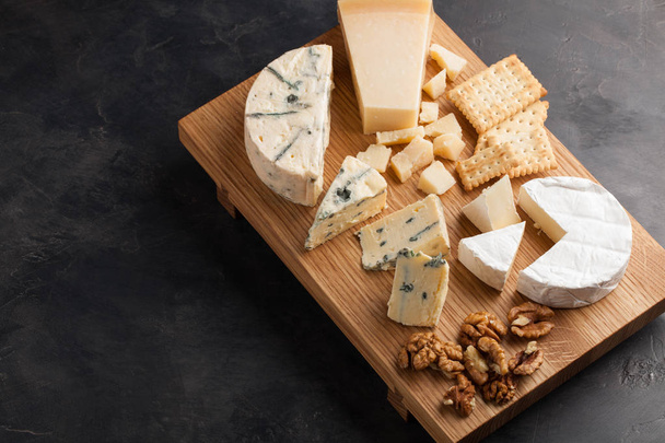 Tasting cheese dish on a wooden plate with walnuts and crackers. Food for wine and romantic, cheese delicatessen on a dark stone table - 写真・画像