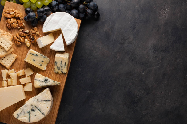 Degustación de plato de queso en un plato de madera. Comida para el vino y romántica, delicatessen queso en una mesa de piedra oscura. Vista superior con espacio de copia
 - Foto, Imagen