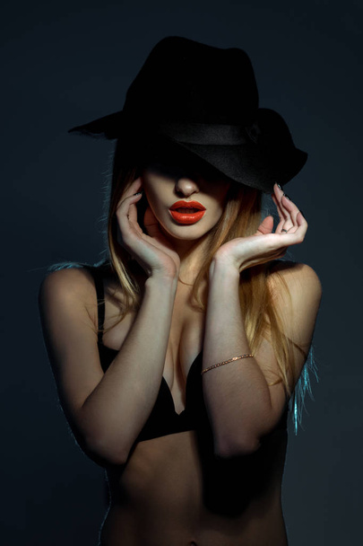 portrait of slim young girl in black bra with red lips wearing a hat with wide brim in studio - Фото, изображение