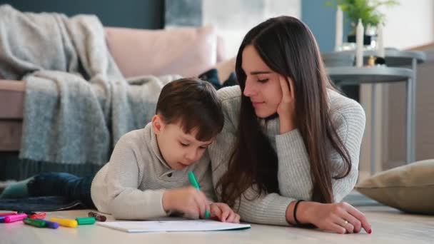 Mooie jonge moeder in een warme trui op de vloer liggen met mijn zoon tekenen met markeringen op papier uitbeelden van zijn familie. Het kind leert om te tekenen. - Video