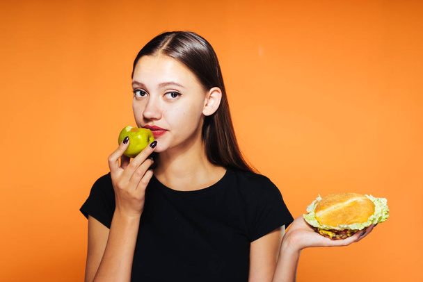 a young sweet girl wants to lose weight, holds a harmful calorie hamburger and a useful apple - Zdjęcie, obraz