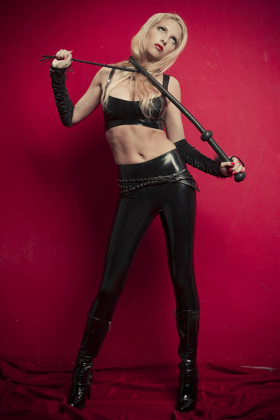 Beautiful blonde mistress posing in studio on a red  with leather whip. Halloween - Foto, Imagem