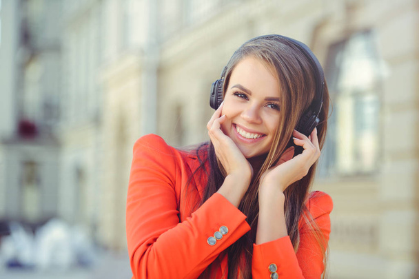 Lachende vrouw luisteren naar muziek op de hoofdtelefoon, kleurrijke rode jas tegen de achtergrond van een Europese stad mode - Foto, afbeelding