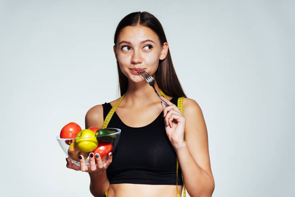 una joven hermosa chica deportiva observa su figura, sostiene un plato con verduras y frutas útiles en sus manos
 - Foto, imagen