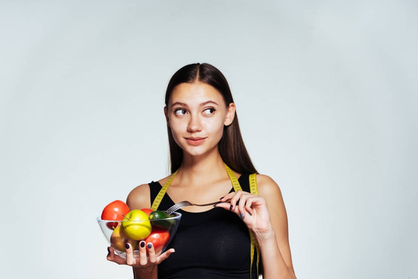 uma jovem bela menina de esportes observa sua figura, segura um prato com vegetais úteis e frutas em suas mãos
 - Foto, Imagem