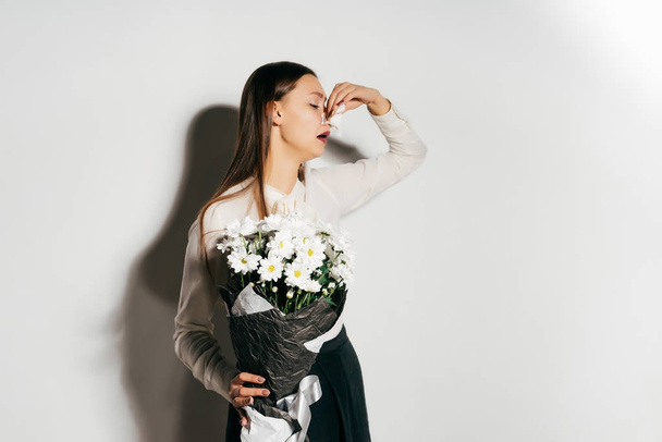 a young beautiful girl in a white shirt is holding a large bouquet of white cet in her hands and sneezes because she is allergic to flowers - Фото, зображення