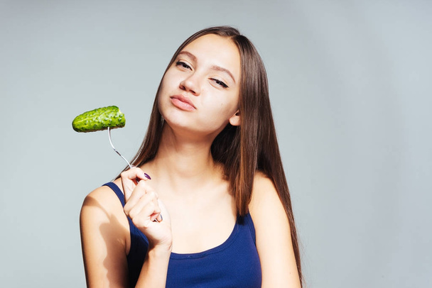 a young beautiful girl in a blue top wants to lose weight, holds a fork with a low-calorie cucumber - Photo, Image