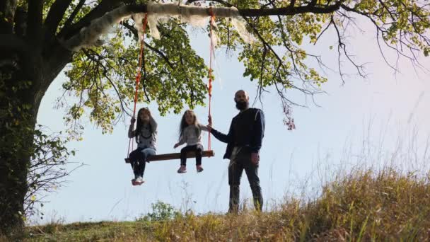 Папа трясет ее дочерьми на качелях под деревом
. - Кадры, видео