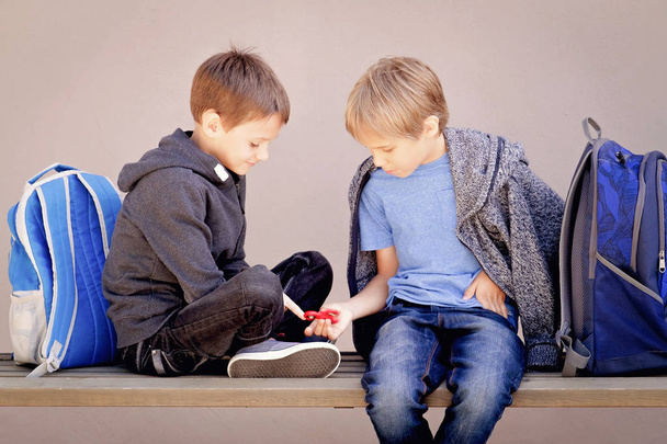 Grundschule, Schule, Freundschaftskonzept - zwei Jungen mit Rucksäcken sitzen, reden und spielen mit Spinner - Foto, Bild