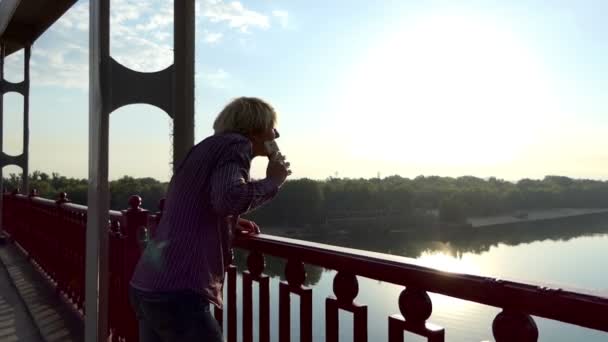 Hombre rubio se para en el puente, llama, levanta sus manos felizmente al atardecer en Slo-Mo
 - Metraje, vídeo