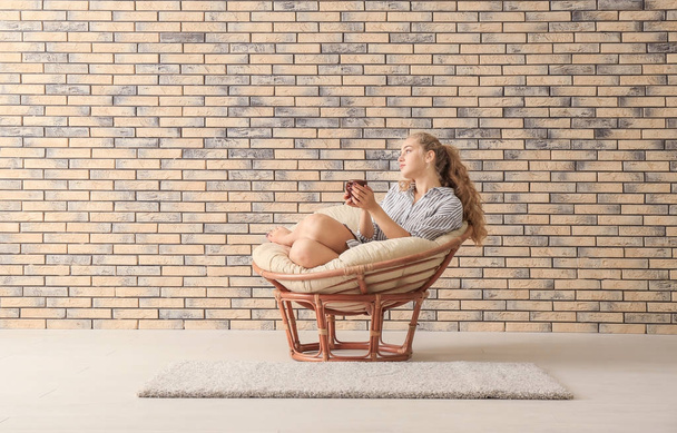 Young woman with cup resting in lounge chair near brick wall - Foto, Bild