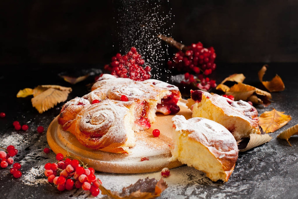 A berry cake, sprinkled with sugar powder, on a round wooden board on a black wooden background surrounded by berries of shoots and autumn leaves - Foto, Imagem