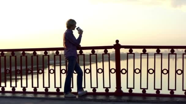 Romantyczny mężczyzna pije kawę z filiżanki papieru i cieszy się zachód słońca na moście w Slo-Mo - Materiał filmowy, wideo