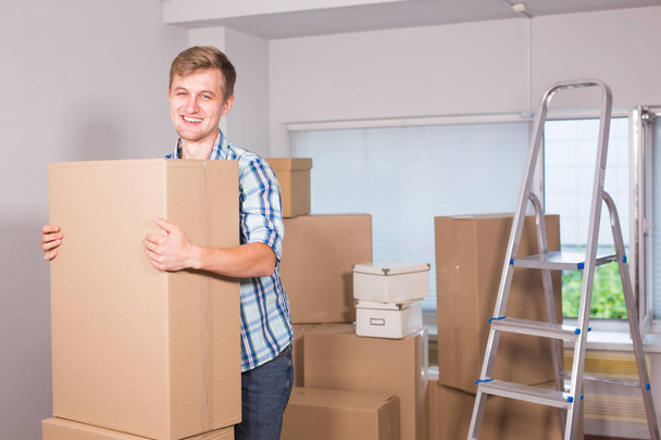 Μετακίνηση σε ένα νέο διαμέρισμα. Χαρούμενος νεαρός άνδρας κρατά ένα χαρτόνι κουτιά και να χαμογελά στη φωτογραφική μηχανή, ενώ άλλα κουτιά από χαρτόνι για τον καθορισμό σε φόντο. - Φωτογραφία, εικόνα