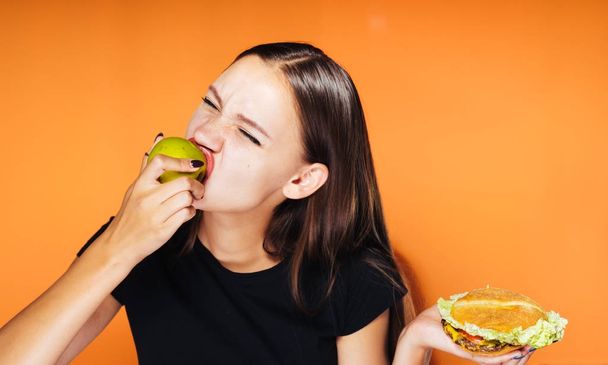 a young beautiful girl wants to lose weight, holds in her hand an unhealthy calorie burger and a useful apple - Photo, image