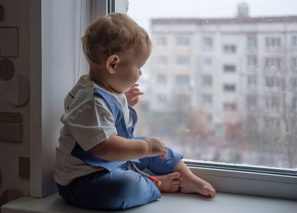 Europejskiego chłopiec siedzi na parapecie i patrząc przez okno - Zdjęcie, obraz