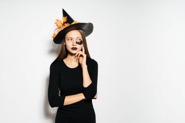 a young sexy gothic girl celebrates halloween, dressed in a witch, on her head a big black hat decorated with yellow autumn leaves - Foto, Imagem