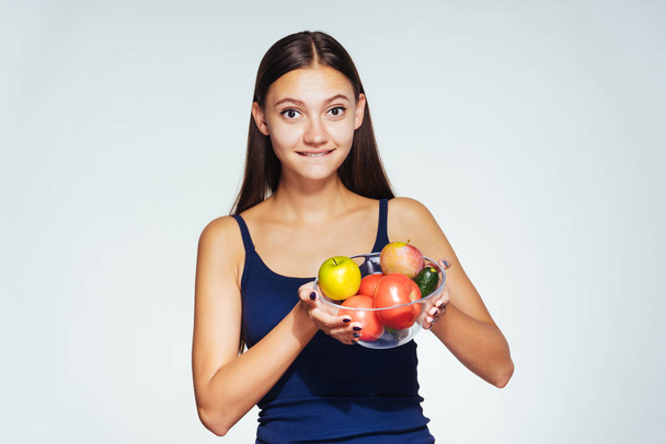 uma jovem atlética em um top azul mantém o controle de sua figura, segura um prato com vegetais úteis e frutas em suas mãos
 - Foto, Imagem