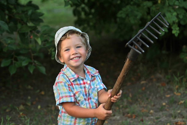El chico de la camisa a cuadros se complace en ayudar a su padre a trabajar en la granja. El hijo de un granjero está de pie con un rastrillo. Niño pequeño con un rastrillo
. - Foto, Imagen