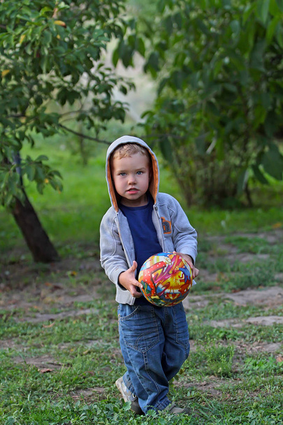 Un niño pequeño en ropa vaquera juega con una bola brillante en el fútbol. Un niño divertido en una capucha sostiene una bola redonda brillante
. - Foto, Imagen