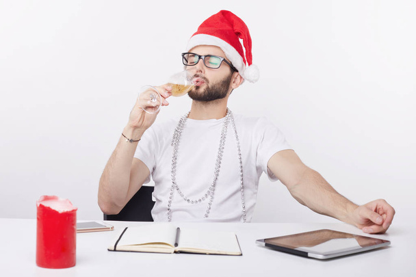 Jonge treurig en eenzaam zakenman vieren van Kerstmis in het kantoor champagne drinken op zijn werkplek. Portret van bebaarde creatieve werknemer overweldigd en verveeld met werk aan vooravond van het nieuwe jaar. - Foto, afbeelding
