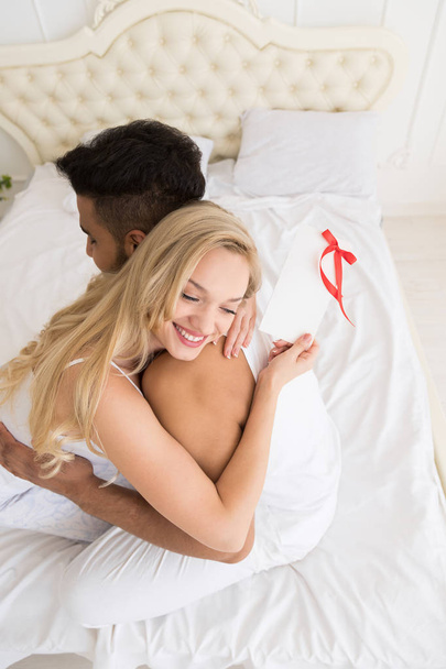 junges glückliches Paar umarmt sich im Bett, hispanischer Mann Frau hält Geschenk-Umschlag mit Schleife, Jubiläumsfeier - Foto, Bild