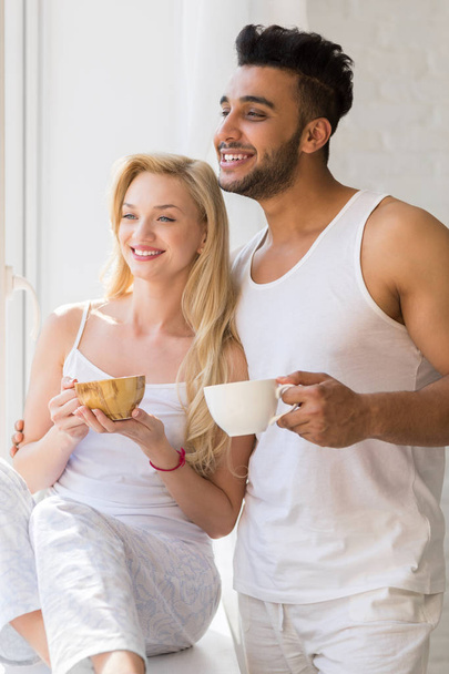 Young Beautiful Couple Stand Near Big Window, Drink Morning Coffee Cup, Happy Smile Hispanic Man Woman - Фото, изображение