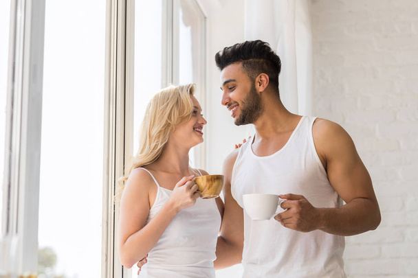 Fiatal gyönyörű pár állni közelében nagy ablak, inni reggel kávéscsésze, boldog mosollyal latin-amerikai férfi nő - Fotó, kép