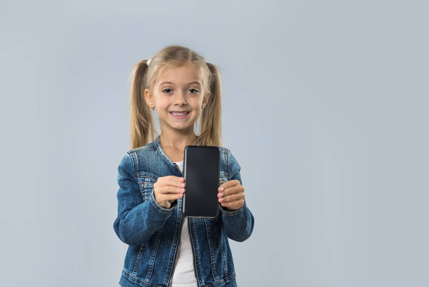 Hermosa niña celebrar celular teléfono inteligente pantalla vacía feliz sonrisa desgaste pantalones vaqueros abrigo aislado
 - Foto, Imagen