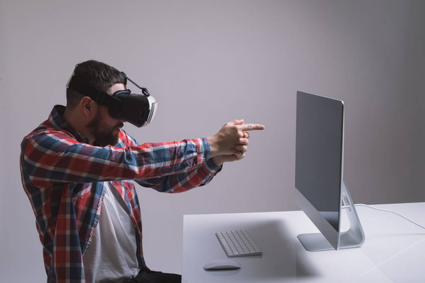 Hombre en casco de realidad virtual juega juego en el escritorio. Man utiliza la pantalla de auriculares VR con auriculares para el juego de realidad virtual en la oficina delante del monitor. Dispositivos de alta tecnología. Creati dispositivo de realidad aumentada
 - Foto, imagen