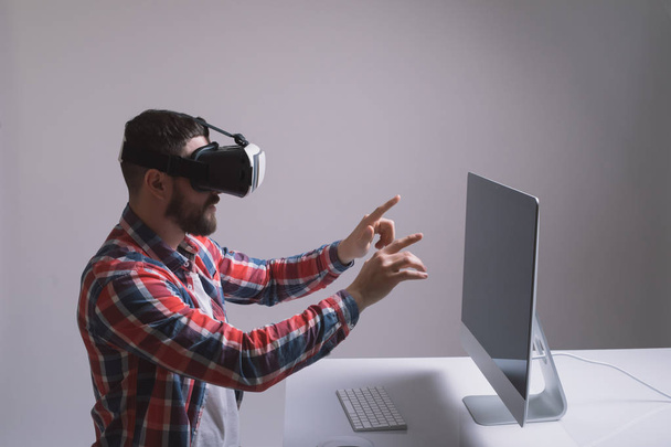 Man in helmet virtual reality plays game at desk. Man uses VR-headset display with headphones for virtual reality game in office in front of monitor. High-tech devices. Augmented reality device creati - Photo, Image
